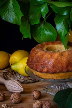 Closeup of lemon bundt on wooden table with lemons, nuts and green leaves, black background, vertical