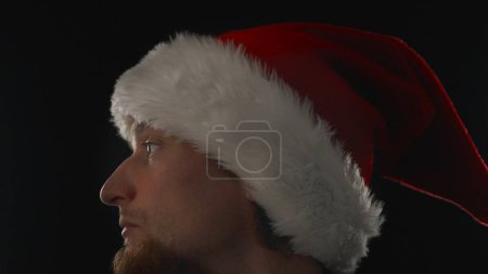 Foto de Un hombre con una barba en un sombrero de Santa sobre un fondo negro, gira la cabeza, de cerca. - Imagen libre de derechos