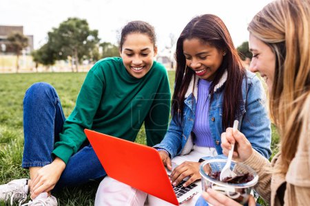 Foto de Jóvenes estudiantes multirraciales trabajando juntas en la computadora portátil durante la pausa para el almuerzo sentadas sobre hierba verde en el campus universitario - Imagen libre de derechos