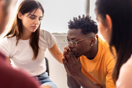 Foto de Support group of people sitting in circle during therapy session sharing personal problems. Men and women supporting each other. Woman supporting african man during a reunion. - Imagen libre de derechos