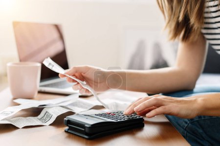 Foto de Young homeowner woman sitting on sofa using calculator and laptop to calculate monthly budget, household expenses, mortgage, rental fees, paying bills online - Imagen libre de derechos