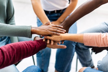 Foto de Grupo joven de personas sentadas en círculo apilando las manos. Amigos multirraciales poniendo sus manos juntas mostrando unidad, apoyo y comunidad. - Imagen libre de derechos