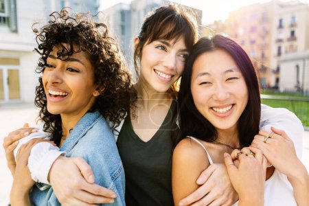 Drei lächelnde junge, unterschiedliche Frauen, die gemeinsam Spaß auf der Straße haben. Glückliche Freundinnen genießen den freien Tag am Wochenende über urbanem Hintergrund. Freundschafts- und Jugendgemeinschaftskonzept.