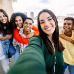 Young group of diverse students having fun together outdoors. Beautiful woman smile at camera taking selfie portrait with diverse friends on the background. Youth community concept.