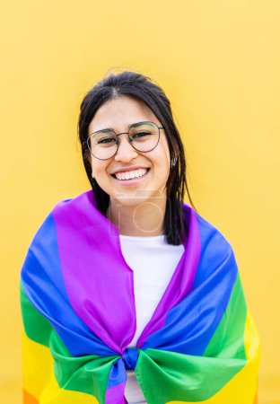 Portrait of young lesbian woman smiling at camera over yellow background celebrating LGBTQ pride day festival covered with rainbow flag proud to be homosexual