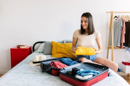 Glückliche junge schöne Frau sitzt auf dem Bett und packt Koffer für die Reise in den Sommerurlaub