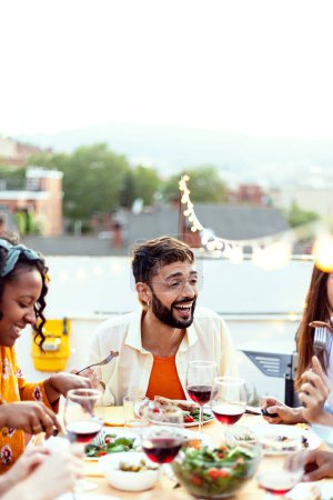Foto vertical de un grupo de jóvenes divirtiéndose brindando por el vino tinto en la cena en la azotea. Amigos multirraciales celebrando juntos en casa terraza durante las vacaciones de verano.