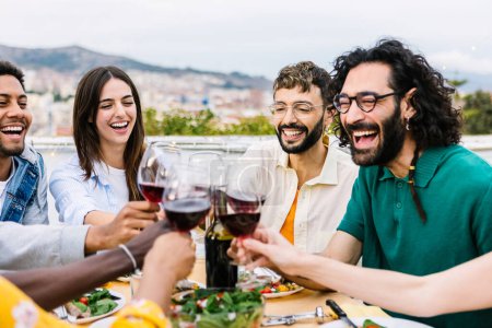 Grupo de jóvenes divirtiéndose brindando por el vino tinto en la fiesta de bb-q en la azotea. Amigos multirraciales celebrando juntos en casa terraza.