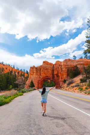 Foto de Joven mujer feliz en el fondo del arco de piedra natural Puente en el Parque Nacional del Cañón Rojo en Utah, EE.UU.. Arco de piedra natural en Red Canyon, Dixie National Forest, Utah, Estados Unidos - Imagen libre de derechos