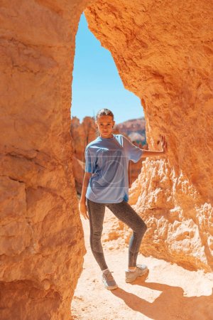 Foto de Senderismo adolescente feliz en Bryce Canyon National Parks, Utah, Estados Unidos. Jovencita parada en un arco de piedra arenisca roja en un sendero - Imagen libre de derechos