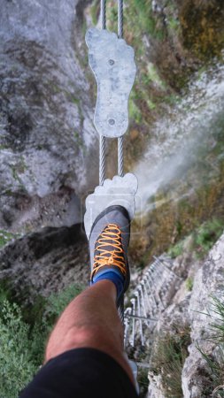 Tourist on foot shaped metal steps, on suspended bridge,over a waterfall, on via ferrata route in Italy, Europe. Summer adventure activities in the Dolomites mountains.