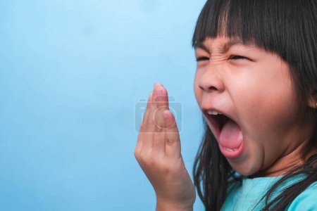 Téléchargez les photos : Petite fille asiatique couvrant sa bouche pour sentir la mauvaise haleine. Enfant fille vérifier la respiration avec ses mains. Problèmes de santé buccodentaire ou concept de soins dentaires. - en image libre de droit
