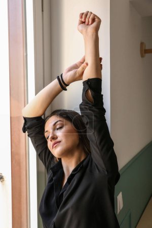 Foto de Joven modelo, vestida con una camisa negra, extendiendo los brazos sobre la cabeza por la ventana, con los ojos cerrados. - Imagen libre de derechos
