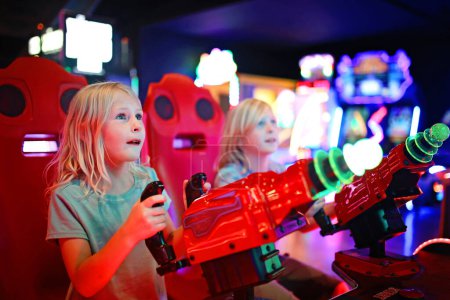 Foto de A little girl and her brother are playing an alien shooting game challenge at a neon light video arcade. - Imagen libre de derechos