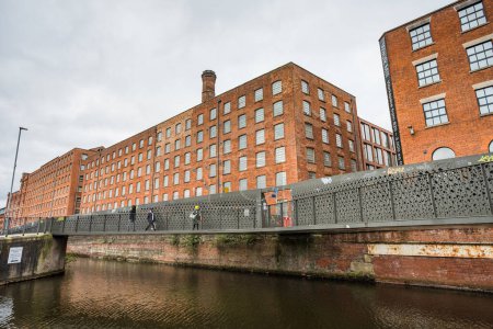 Téléchargez les photos : Incidental people walking over footbridges across Rochdale Canal where it meets the New Islington marina in Manchester seen in February 2023.  The grade II former cotton mill known as Beehive Mill dominates the background. - en image libre de droit