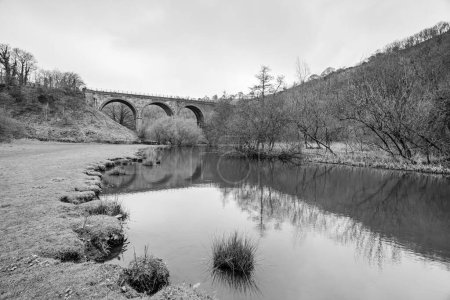 A black and white image of the Monsal Head Bridge reflecting in the River Wye pictured under a cloudy sky.