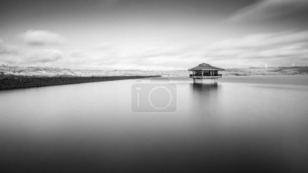 A black and white long exposure of the valve tower at Carsington Water in Derbyshire pictured under moving clouds.