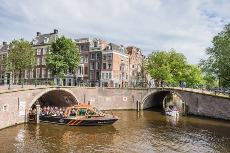 Téléchargez les photos : Un petit bateau passe sous le premier de sept ponts qui enjambent le canal Reguliersgracht à Amsterdam, aux Pays-Bas vu le 27 mai 2024 comme un bateau de croisière parcourt le canal Herengracht. - en image libre de droit
