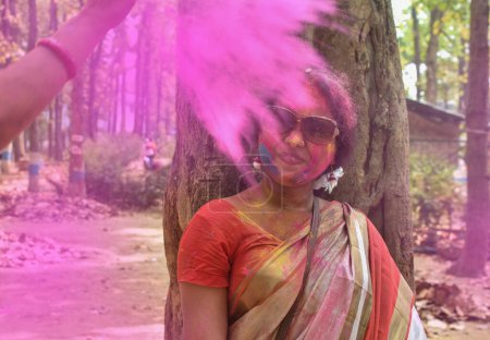 Spritzende trockene organische Farbe oder Farbe oder Gulal oder Bier oder Holi-Puder auf eine schöne junge indische Frau, die Sari auf Holi oder dem Fest der Farben trägt. Bengalische Frauen spielen oder feiern oder genießen das traditionelle Fest.
