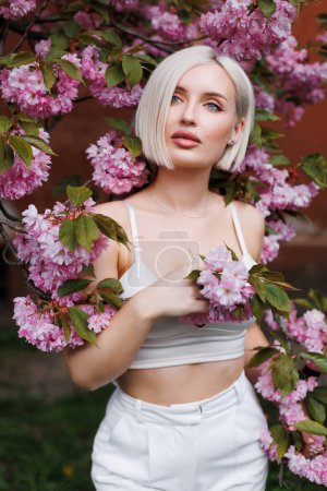 Photo for Sexy woman with short white hair in the sunlight on a beige background and a branch of blooming sakura. - Royalty Free Image