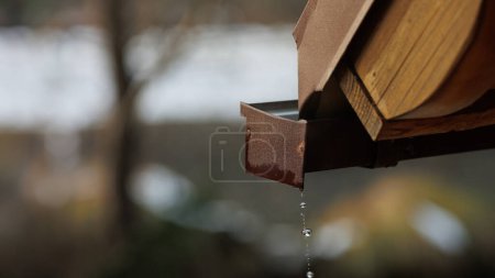 A macro view captures water droplets dripping from the cottage gutter against a spring snowmelt backdrop