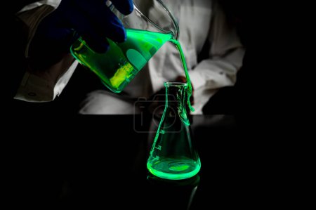 Un chercheur scientifique expérimentant une solution fluorescente verte dans une fiole conique en verre dans un laboratoire biomédical sombre pour le développement de la médecine de soins de santé. Espace de copie fond noir