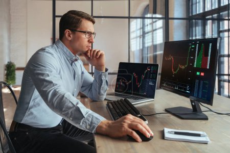 Photo for Side view of crypto broker sitting at office table, analyzing market, looking at screen of personal computer with candlestick chart, touching chin with hand, thinking, wearing shirt and glasses - Royalty Free Image