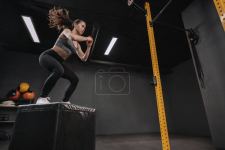 Téléchargez les photos : Femme faisant des exercices de saut en boîte dans le cadre de son entraînement Crossfit. Athlète féminine faisant des squats et sautant sur la boîte dans le gymnase d'entraînement sombre. Espace de copie - en image libre de droit