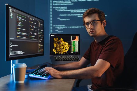Portrait of Intelligent male programmer working on pc writing brand new code at his home office, looking at camera on background with digital wall with application info page. Data science concept