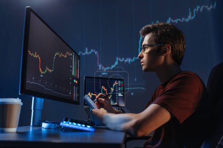 Photo for Side view of young man trader analyst at working table, analyzing financial chart, stock quotes, share prices, trading online, checking data on cryptocurrency graph on computer screen, making notes - Royalty Free Image