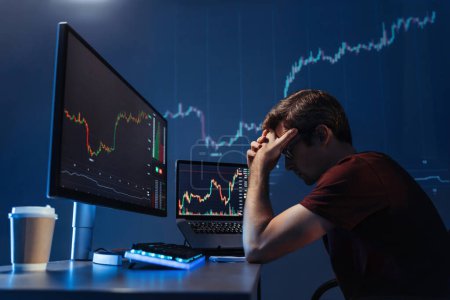 Seitenansicht eines deprimierten nachdenklichen männlichen Krypto-Anlegers, der den Kopf in den Händen vor dem Computer mit Kerzenständer-Chart des Krypto-Devisenmarktes hält, versagt Geld zu verdienen, verärgert über die globale Rezession