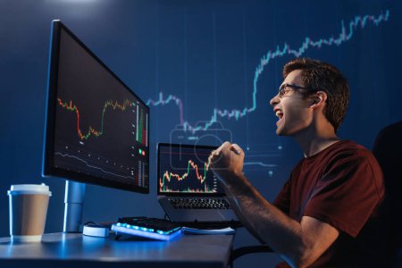 Photo for Side view of male crypto trader doing yes gesture shouting with triumph. Exited man looking at screen with digital candlestick chart of cryptocurrency trading and investments market - Royalty Free Image