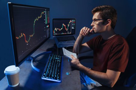 Téléchargez les photos : Crypto investor working on pc, noting down results of his trading market analysis, sitting at table, looking at display with concentration, analyzing price movements, adjusting investment strategy - en image libre de droit