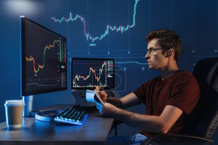 Side view of male crypto broker sitting at his workplace at night, blue digital wall with financial diagram background. Trader checking candlestick chart, making notes. Cryptocurrency