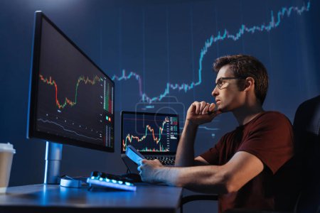 Téléchargez les photos : Side view of concentrated crypto trader sitting in front of computers, making professional analysis of candlestick chart, creating strategy, holding pen and notepad, looking at monitor, touching chin - en image libre de droit