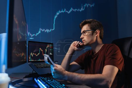 Téléchargez les photos : Crypto trader freelancer sitting at table with notepad late night. Noting down info after checking ico and trading market dynamics on his personal computer with candlestick chart on display - en image libre de droit