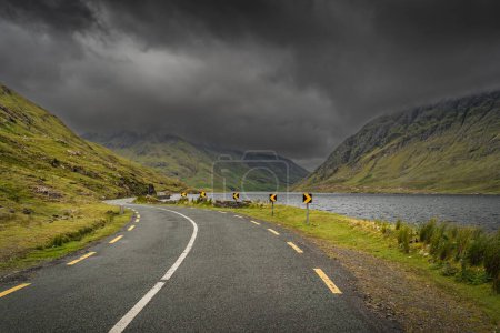 Photo for Winding road leading trough Doloough Valley with lake, between Glenummera and Glencullin mountain ranges with dark dramatic sky, Mayo, Ireland - Royalty Free Image