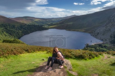 Foto de Madre e hija, familia, sentadas en una gran roca y sonriendo. Lough Tay, llamado Guiness Lake en Wicklow Mountains, Irlanda - Imagen libre de derechos
