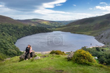 Foto de Madre e hija, familia, sentadas en una gran roca en el borde del acantilado. Lough Tay, llamado Guiness Lake en Wicklow Mountains, Irlanda - Imagen libre de derechos