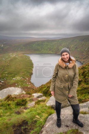 Foto de Mujer de pie sobre la roca en la cima de la montaña y sonriente, lago, Lough Bray, abajo en el fondo. Senderismo en Wicklow Mountains, Irlanda - Imagen libre de derechos