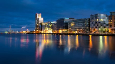 Photo for Modern offices and apartment buildings on Sir John Rogerson Quay and Poolbeg Powerstation chimneys, blurred Liffey River at dusk, Dublin, Ireland - Royalty Free Image