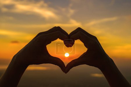 Woman hands silhouette on beautiful sunset above the mountain in the form of love heart-shaped hand gesture Concept of love, life, romance.