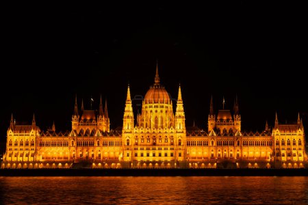 Esta impresionante fotografía captura el esplendor nocturno del Orszaghaz, el icónico edificio del Parlamento de Budapest, que se encuentra magníficamente iluminado a lo largo del río Danubio. La imagen