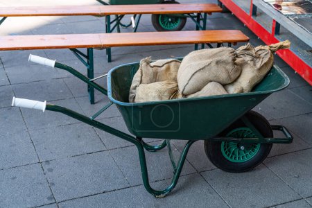 Photo for Green wheelbarrow with sacks stands near wooden benches. - Royalty Free Image