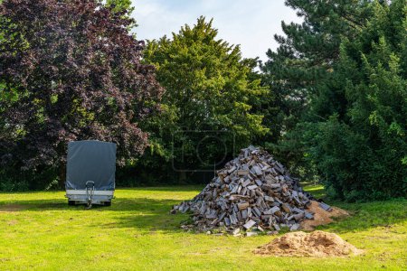 Photo for A large pile of sawn firewood and a car trailer on a green lawn against the backdrop of large trees. - Royalty Free Image