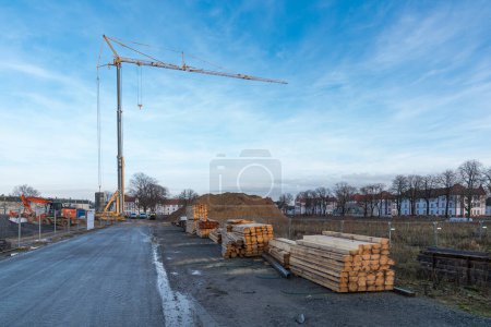 Foto de Torre grúa y excavadora en un sitio de construcción con madera apilada. Cielo azul con nubes claras - Imagen libre de derechos