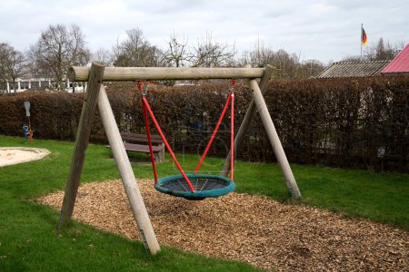 Wooden swing on a small playground with green grass. Wooden bench in the background.