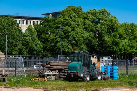 Pequeño tractor de construcción y materiales de construcción apilados cerca de una valla metálica en un paisaje de la ciudad.