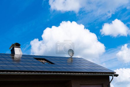 The roof of the building with a chimney and a satellite dish is covered with solar panels. Blue sky with beautiful white clouds.