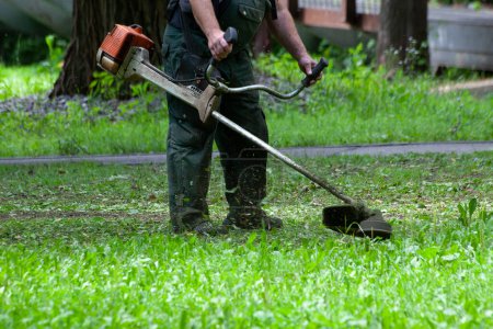 Arbeiter mähen Gras mit Benzin-Rasentrimmer im Stadtpark oder Hinterhof. Werkzeuge und Geräte für die Gartenpflege. Prozess des Rasentrimmens mit dem Handmäher.